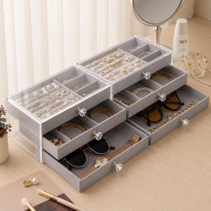 May include: A clear acrylic jewellery organiser with multiple pull-out drawers lined in soft grey velvet. The drawers feature crystal-style knobs and contain various accessories including gold-tone bracelets, necklaces, sunglasses, and a wristwatch.
