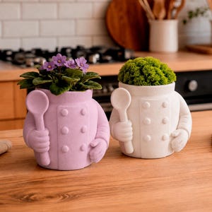 Op de afbeelding: Twee keramische plantenpotten in de vorm van een koksbuis, één lichtpaars en één wit. Elke pot heeft gebeeldhouwde knopen, armen en een hand die een lepel vasthoudt. De paarse pot bevat een bloeiende plant, de witte bevat groen mos.