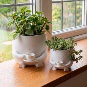 May include: Two matte white ceramic planters shaped like small figures with arms and legs. Each planter features a smiling face and holds a tiny cup and saucer. The larger planter contains a green leafy plant, while the smaller one holds a succulent.