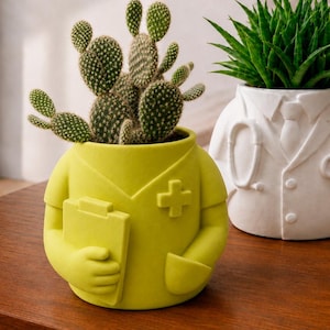 Può includere: Due vasi in ceramica a forma di professionisti medici. Uno è verde lime, a forma di divisa con cartellina e croce, contenente un cactus. L'altro è bianco, a forma di camice da laboratorio con stetoscopio, contenente una pianta succulenta.