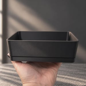 May include: A matte black rectangular container with a smooth finish and slightly tapered sides. The vessel features a clean, minimalist design with a distinct horizontal seam near the base, providing a structured and uniform appearance.