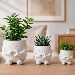 May include: Three white ceramic planters in descending sizes, each shaped like a small figure with arms forming a heart shape over its chest and two feet at the base. The planters hold a leafy green plant, a spiky succulent, and a small green herb.