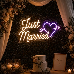 May include: A neon sign featuring the text Just Married in a warm white script font, accompanied by a purple heart pierced by an arrow. The sign is mounted on a dark green foliage wall, positioned above a two-tiered white cake and a framed sign.