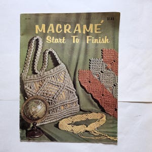 May include: A vintage instructional booklet titled MACRAMÉ Start To Finish featuring a beige macramé handbag, a matching belt, and decorative woven panels. A small globe sits in the lower left corner against a dark green draped background.