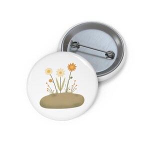 May include: A round pinback button featuring a white background with a graphic illustration of three stylized flowers in shades of yellow and orange growing from a solid tan mound. The back of the button is silver metal with a pin mechanism.