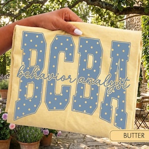 May include: A butter yellow t-shirt featuring the large blue block letters BCBA with a white polka dot pattern. Overlaid across the centre is the text behaviour analyst in a blue cursive script font. The shirt is held up against an outdoor background.