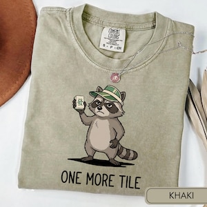 Könnte beinhalten: Ein khakifarbenes Kurzarm-T-Shirt mit einer Grafik eines Waschbären, der einen grün-weiß gestreiften Hut trägt und einen Mahjong-Stein hält. Unter dem Waschbären steht der Text ONE MORE TILE in schwarzen Großbuchstaben. Das Shirt hat einen Rundhalsausschnitt.