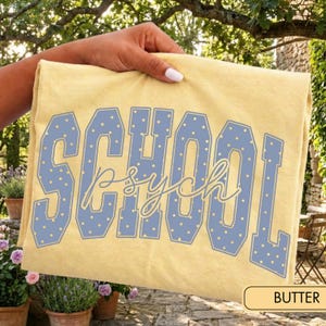 May include: A pale yellow t-shirt featuring the word SCHOOL in large, blue, polka-dotted block letters. Overlaid across the center is the word psych written in a thin, cursive, light yellow script font. The design is centered on the fabric.