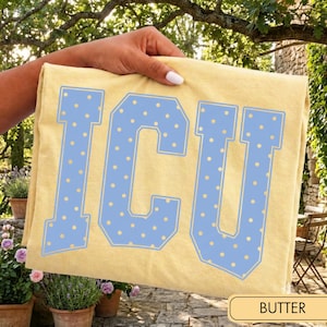 May include: A pale yellow t-shirt featuring the letters ICU in a large, block-style font. The letters are light blue with a repeating pattern of small yellow dots and a thin light blue outline, creating a bold, collegiate-inspired graphic design.