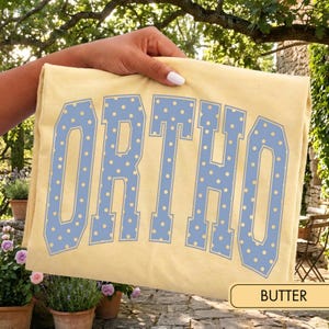 May include: A pale yellow t-shirt featuring the word ORTHO printed in large, block-style letters. The letters are light blue with a repeating pattern of small, pale yellow polka dots throughout the text. The fabric has a smooth, matte finish.