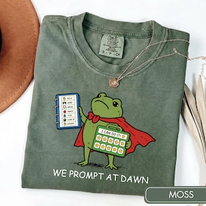 May include: An olive green t-shirt featuring a cartoon frog wearing a red cape. The frog holds a green chart with the text I CAN DO IT and ten yellow stars. Next to the frog is a blue checklist with icons and text. The text WE PROMPT AT DAWN is printed below.
