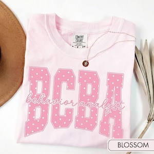 May include: A light pink short-sleeve t-shirt with a Comfort Colors brand label at the neck. The front features large pink block letters with white polka dots spelling BCBA, overlaid with the cursive white text behaviour analyst.
