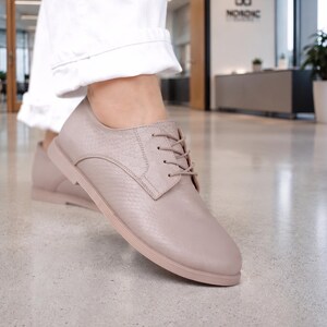 Könnte beinhalten: Ein Paar helltaupefarbene Schnürschuhe mit einem dezenten, schlangenlederartigen Strukturmuster auf dem Obermaterial. Die Schuhe verfügen über farblich abgestimmte Schnürsenkel, eine abgerundete Schuhspitze und eine flache, tonale Sohle.