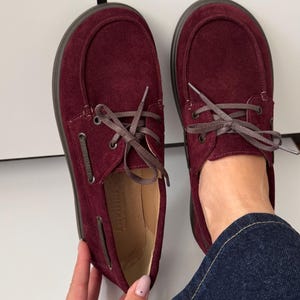 Könnte beinhalten: Ein Paar tiefburgunderrote Wildlederschuhe mit abgerundeter Zehenpartie, einer mokassinartigen Naht über dem Vorderblatt und dunkelbraunen Schnürsenkeln durch Metallösen. Die Schuhe haben eine flache, dunkelbraune Sohle und ein beiges Innenfutter.