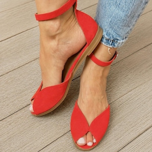 May include: A pair of bright red flat sandals featuring a peep-toe design and an ankle strap with a buckle closure. The sandals have a soft, matte texture and a simple, low-profile sole, providing a clean look for casual footwear options.