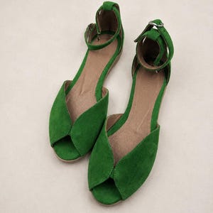 May include: A pair of vibrant green suede flat sandals featuring an open-toe design, a curved vamp, and an adjustable ankle strap with a silver-toned metal buckle. The interior footbed is a neutral tan color, and the soles are flat and low-profile.