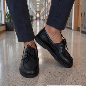 Könnte beinhalten: Ein Paar schwarze Bootsschuhe aus Leder mit Mokassin-Zehenpartie, Schnürung mit schwarzen Schnürsenkeln und einer dicken schwarzen Gummisohle. Die Schuhe haben eine glatte Oberfläche und werden zu einer dunkelgrauen Hose getragen.