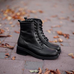 May include: A pair of black leather ankle boots featuring a textured finish, rounded toes, and black laces that extend up the front. The boots have a side zipper closure, a pull tab at the heel, and a low, block heel with a thick, durable rubber sole.