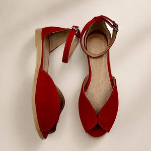 May include: A pair of red flat sandals with a peep-toe design, featuring a closed heel and an adjustable ankle strap with a silver-toned metal buckle. The interior lining is a light beige color, and the soles are a flat, light tan material.