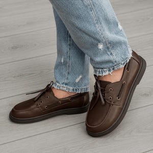 Könnte beinhalten: Ein Paar dunkelbraune Bootsschuhe aus Leder mit Mokassin-Zehenpartie, passenden braunen Schnürsenkeln und metallischen Ösen. Die Schuhe werden zu einer hellblauen, abgenutzten Jeans mit ausgefranstem Saum am Knöchel getragen.