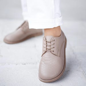 May include: A pair of taupe-colored lace-up oxford shoes featuring a textured snakeskin-like embossed pattern on the upper material. The shoes have a rounded toe, matching taupe laces, and a flat sole, worn with white cuffed trousers.