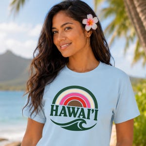 May include: A light blue short-sleeved t-shirt featuring a circular graphic with a multi-colored rainbow arch, the text HAWAI'I in dark green capital letters, and a dark green wave design at the bottom of the circular emblem.