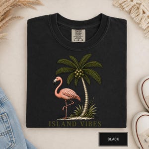 May include: A black t-shirt featuring a graphic of a pink flamingo standing next to a palm tree with coconuts. The text "ISLAND VIBES" is printed in a muted green font below the graphic. The shirt has a Comfort Colors label showing size L-G.