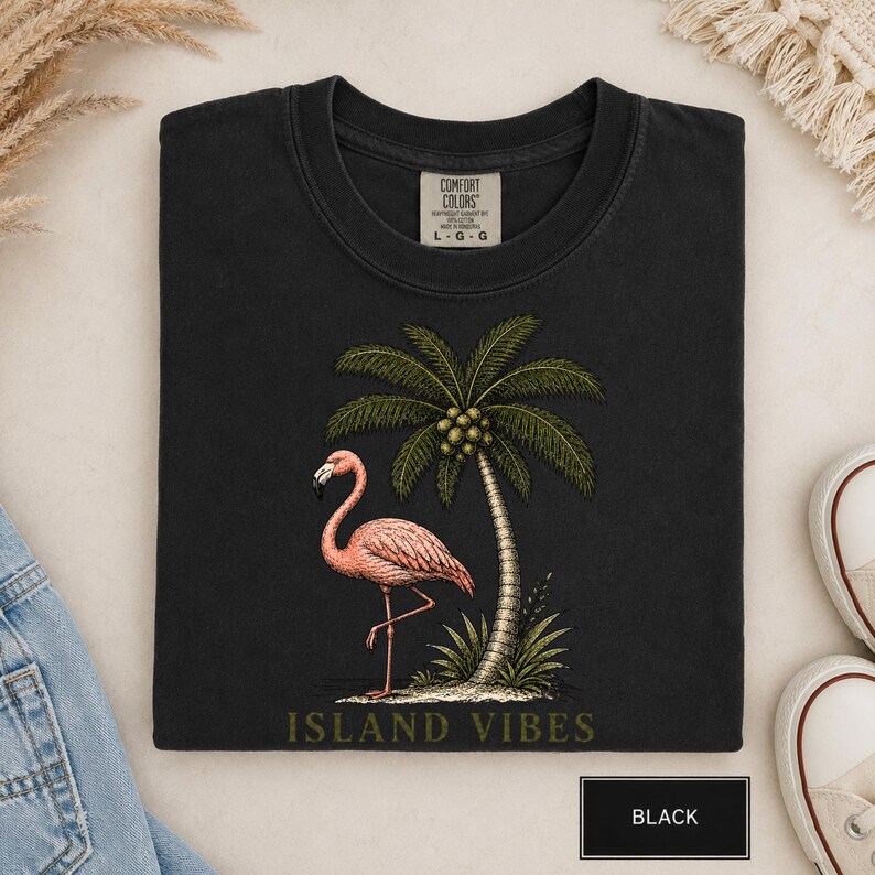May include: A black t-shirt featuring a graphic of a pink flamingo standing next to a palm tree with coconuts. The text "ISLAND VIBES" is printed in a muted green font below the graphic. The shirt has a Comfort Colors label showing size L-G.
