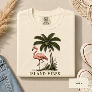May include: An ivory t-shirt featuring a graphic print of a pink flamingo standing next to a green palm tree. The text "ISLAND VIBES" is printed in dark green below the illustration. The shirt has a Comfort Colors brand label at the neck.