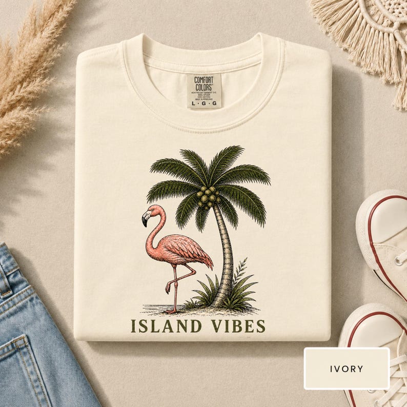 May include: An ivory t-shirt featuring a graphic print of a pink flamingo standing next to a green palm tree. The text "ISLAND VIBES" is printed in dark green below the illustration. The shirt has a Comfort Colors brand label at the neck.