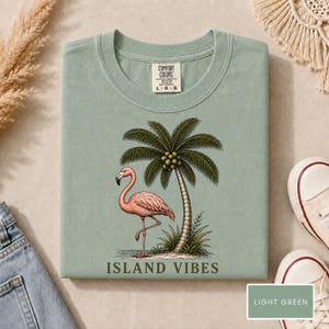 May include: A sage green t-shirt featuring a graphic print of a pink flamingo standing next to a palm tree. The text "ISLAND VIBES" is printed in dark green below the illustration. The shirt has a Comfort Colors brand label at the neck.