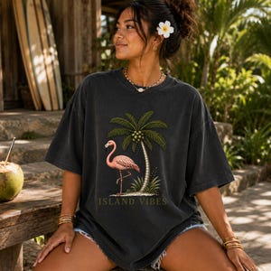 May include: A charcoal grey oversized t-shirt featuring a graphic print of a pink flamingo standing next to a palm tree with coconuts. The text ISLAND VIBES is printed in a muted gold font below the graphic. The shirt has a crew neckline.