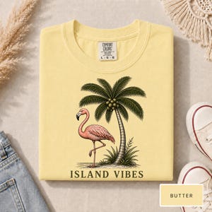 May include: A butter yellow t-shirt featuring a graphic of a pink flamingo standing next to a palm tree. The text ISLAND VIBES is printed in black below the graphic. The shirt has a Comfort Colors brand label at the neck with size L G G.