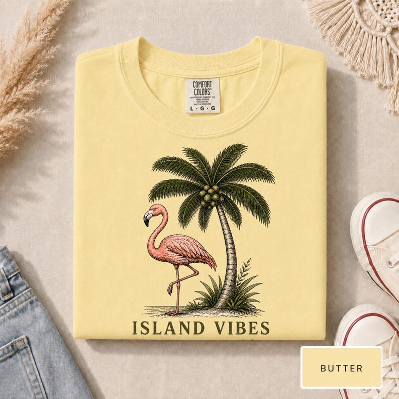 May include: A butter yellow t-shirt featuring a graphic of a pink flamingo standing next to a palm tree. The text ISLAND VIBES is printed in black below the graphic. The shirt has a Comfort Colors brand label at the neck with size L G G.