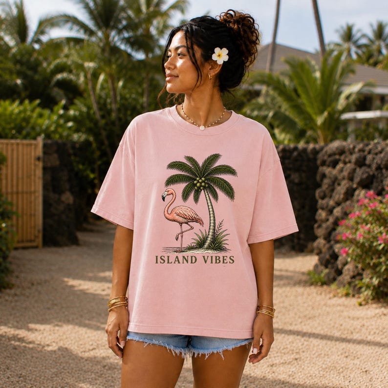 May include: A light pink short-sleeved t-shirt featuring a graphic print of a pink flamingo standing under a green palm tree. The text ISLAND VIBES is printed in dark brown capital letters below the graphic illustration on the front of the shirt.
