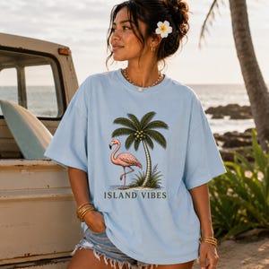 May include: A light blue t-shirt featuring a graphic print of a pink flamingo standing next to a palm tree. The text ISLAND VIBES is printed in dark green letters below the illustration. The shirt has a crew neckline and short sleeves.