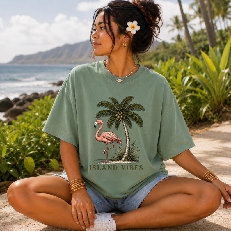 May include: A sage green short-sleeve t-shirt featuring a graphic print of a pink flamingo standing next to a palm tree with the text ISLAND VIBES printed in dark green lettering below the illustration, centred on the front of the garment.