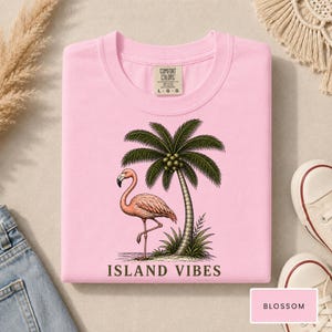 May include: A light pink t-shirt featuring a graphic print of a flamingo standing next to a palm tree. The text "ISLAND VIBES" is printed in dark brown letters below the graphic. The shirt has a Comfort Colors brand label showing size L.