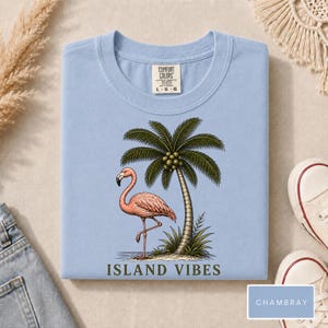 May include: A light blue crew neck t-shirt featuring a graphic of a pink flamingo standing next to a palm tree. The text "ISLAND VIBES" is printed in dark green below the graphic. The shirt has a Comfort Colors brand label at the neck.
