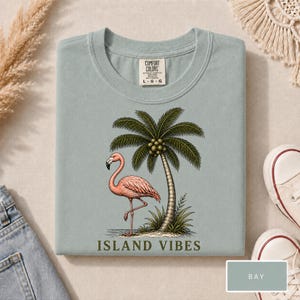 May include: A light blue t-shirt featuring a graphic of a pink flamingo standing next to a palm tree. The text ISLAND VIBES is printed in dark green below the illustration. The shirt has a Comfort Colors brand label at the neck showing size L.