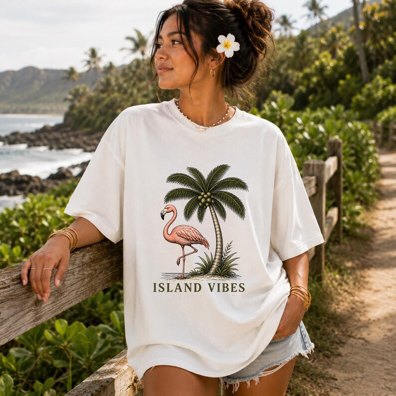 May include: An off-white short-sleeve t-shirt featuring a graphic print of a pink flamingo standing next to a green palm tree. The text ISLAND VIBES is printed in dark green capital letters below the illustration on the front of the shirt.