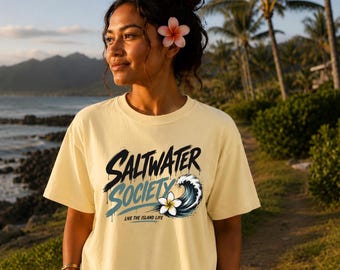 May include: A pale yellow short-sleeve t-shirt featuring a graphic design with the text "SALTWATER SOCIETY" in black and blue, "LIVE THE ISLAND LIFE" in black, and an illustration of a crashing ocean wave with a white and yellow plumeria flower.