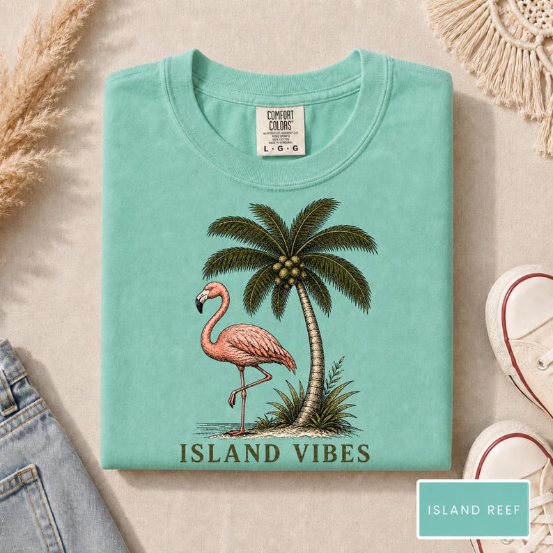 May include: A light teal t-shirt featuring a graphic of a pink flamingo standing next to a palm tree. The text "ISLAND VIBES" is printed in dark green below the illustration. The shirt has a Comfort Colors brand label at the neck.