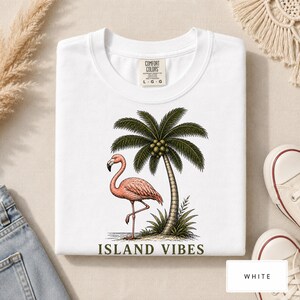 May include: A white t-shirt featuring a graphic print of a pink flamingo standing on one leg next to a palm tree with coconuts. The text "ISLAND VIBES" is printed in dark green capital letters below the illustration. The shirt has a Comfort Colors label.
