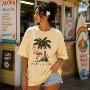 May include: A pale yellow short-sleeve t-shirt featuring a graphic print of a pink flamingo standing next to a palm tree with the text ISLAND VIBES printed below the illustration. The shirt has a crew neckline and a relaxed, loose-fitting silhouette.