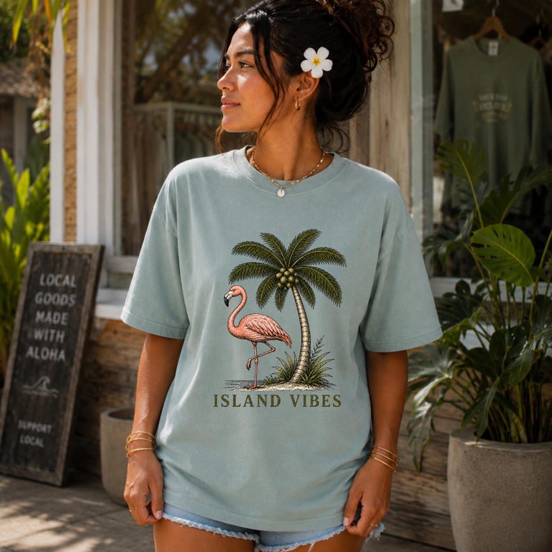 May include: A light sage green short-sleeve t-shirt featuring a graphic print of a pink flamingo standing next to a palm tree. The text ISLAND VIBES is printed in dark green letters below the graphic. The shirt has a crew neckline and a relaxed fit.