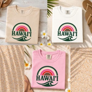May include: Three short-sleeve t-shirts in beige, white, and light pink. Each shirt features a circular graphic with a rainbow in shades of brown, red, and pink, a dark green wave, and the text HAWAI'I in dark green capital letters.