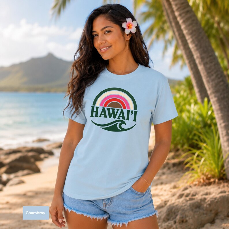 May include: A light blue short-sleeved t-shirt featuring a circular graphic with a multi-colored rainbow arch, the text HAWAI'I in dark green capital letters, and a dark green wave design at the bottom of the circular emblem.