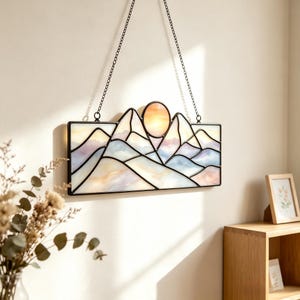 Peut inclure: Un attrape-soleil rectangulaire en vitrail représentant un paysage de montagne avec un soleil orange circulaire. L'objet est composé de lignes de plomb en métal noir et contient des panneaux de verre blanc, bleu pâle et violet doux, suspendu par deux chaînes.