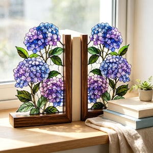 以下が含まれることがあります： 青と紫のアジサイの花の房と緑の葉が描かれたステンドグラスパネルを備えたブックエンドのペア。ダークブラウンの木製ベースに固定されています。ガラスパネルには、暗い鉛のラインで縁取られた複雑な花の模様が施されています。